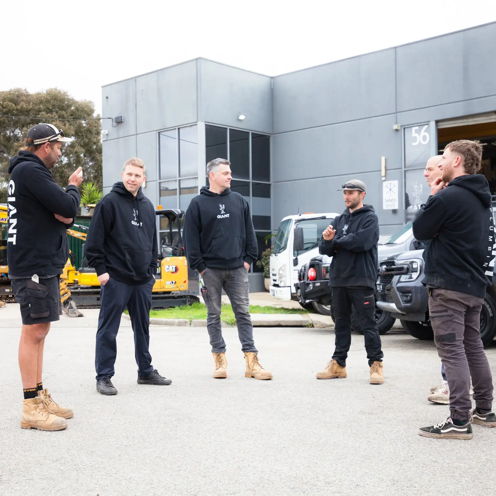 The five Giant Plumbing team members talking in the yard outside the Coburg North workshop, their excavators, tip truck and utes behind them