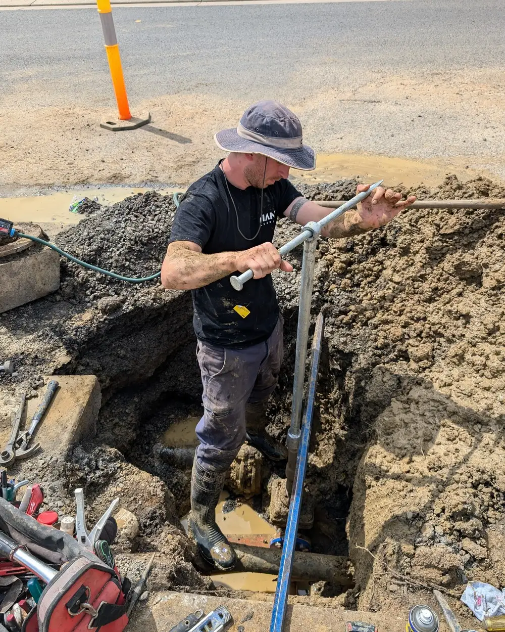 A Giant Plumbing worker in an excavation beside a commercial street, valve key in hand