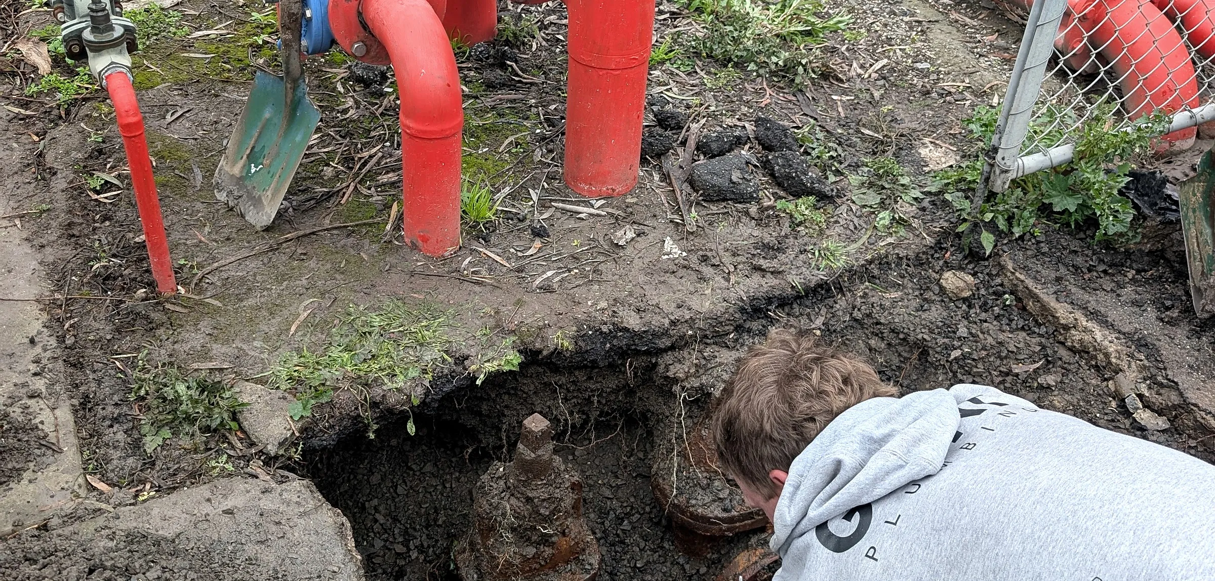 A Giant Plumbing team member in the excavation beside a red fire booster assembly, the old hydrant valve exposed below it