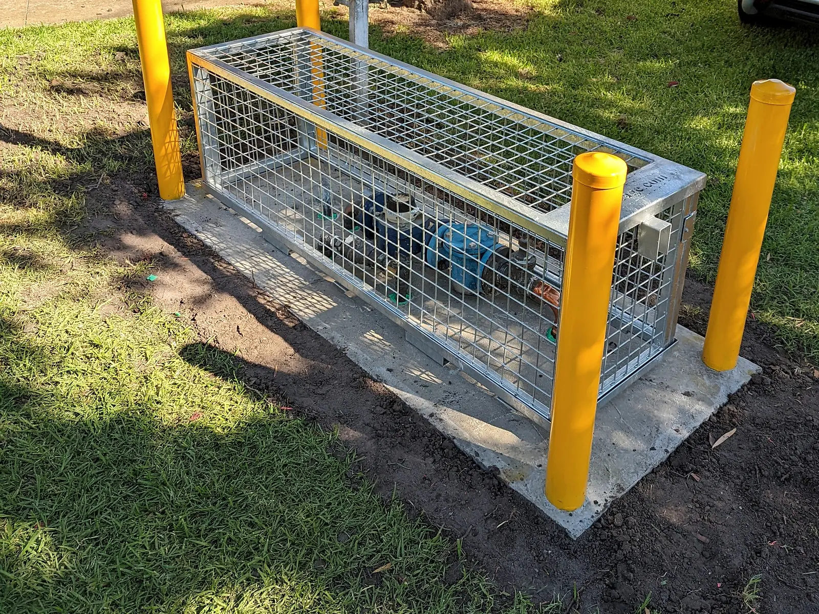 A backflow assembly in a galvanised cage on a concrete pad behind yellow bollards