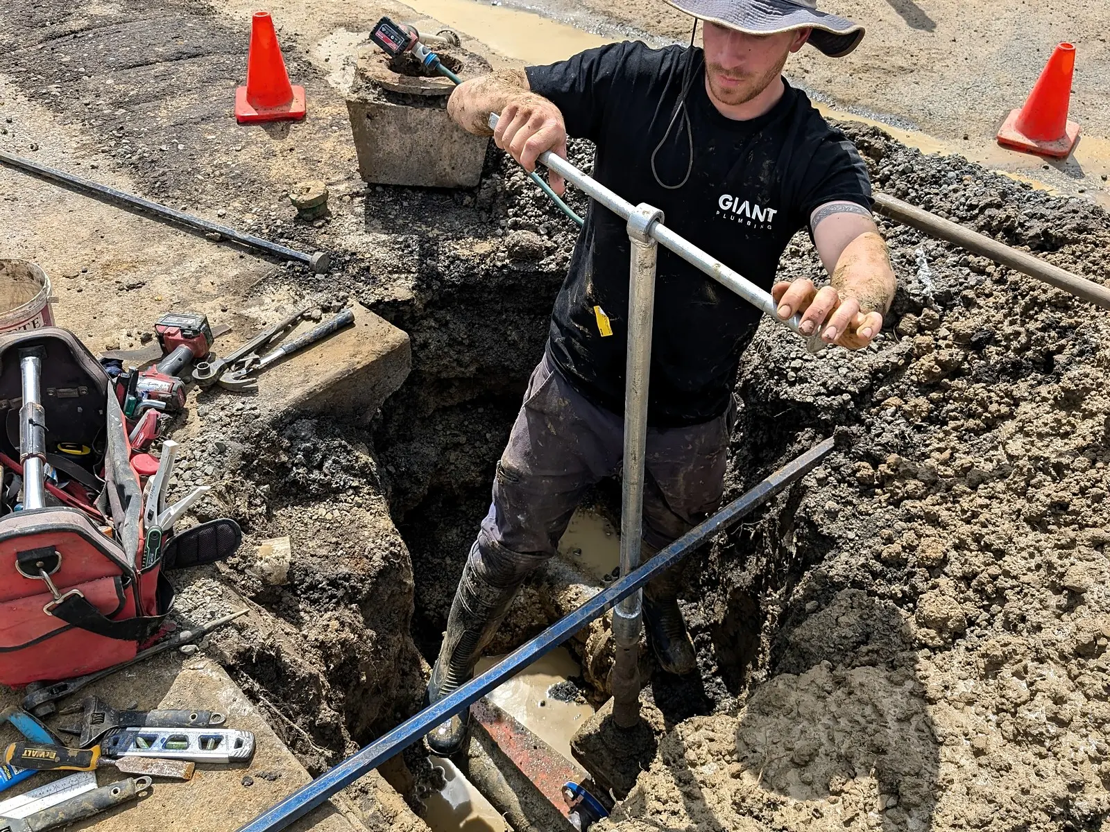 A Giant Plumbing team member turns a valve key in a footpath excavation, with a red pipe in the water below