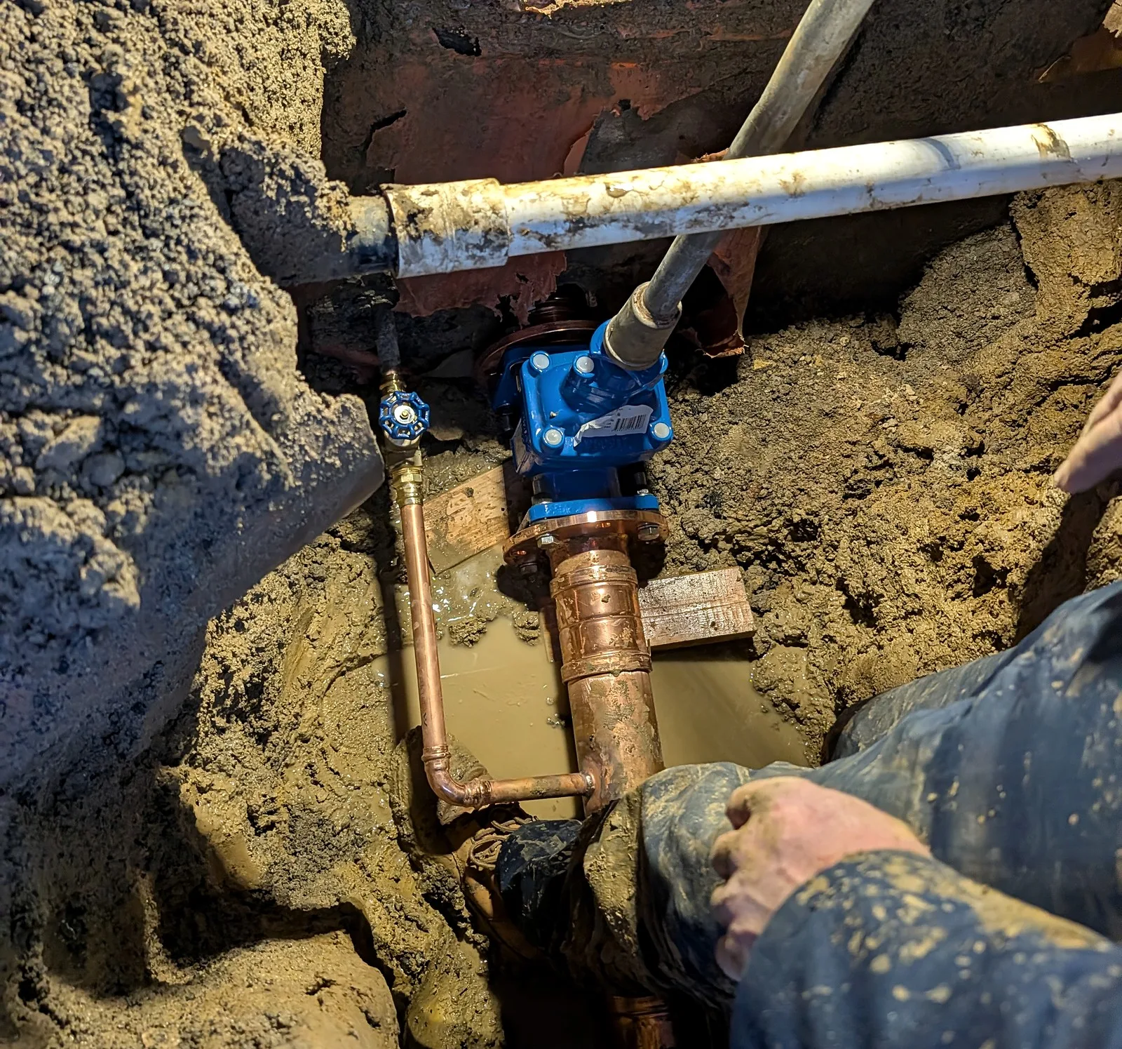 Hands fit a new blue isolation valve to a copper fire service riser deep in an excavation