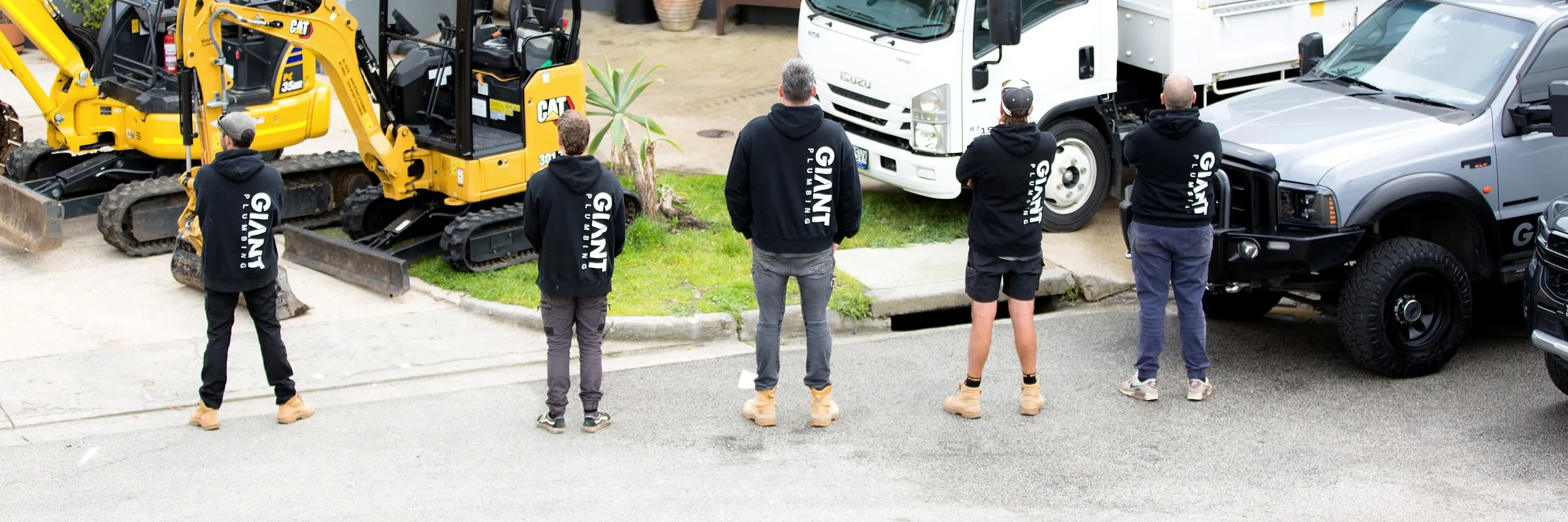 The five Giant Plumbing team members from behind, GIANT PLUMBING across their hoodies, facing the tip truck and excavators at the Coburg North yard