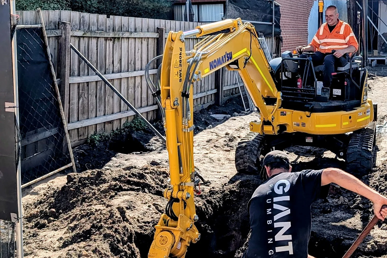 A Komatsu mini excavator digs between fences in a backyard while a Giant Plumbing team member digs by hand