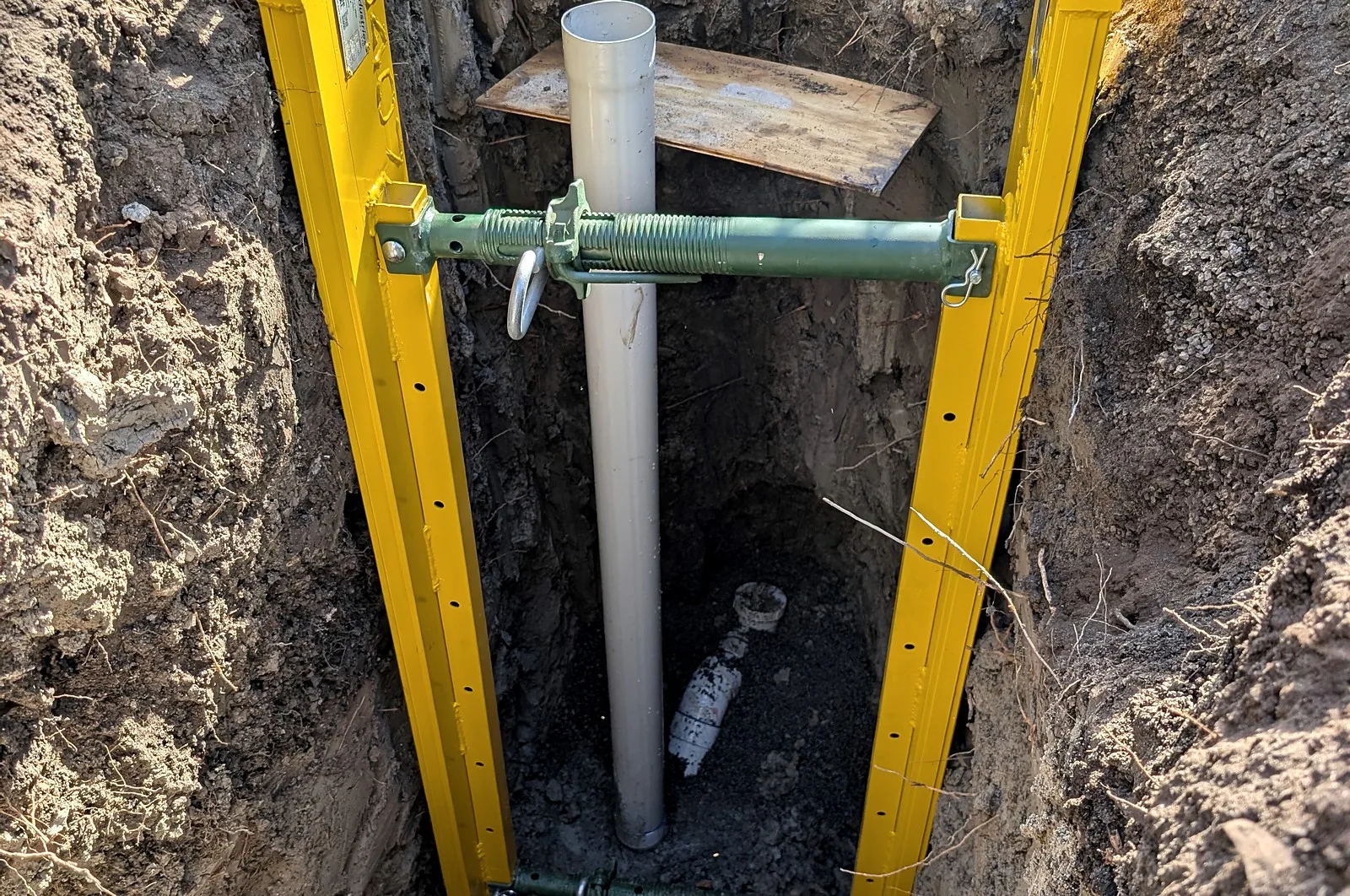 A deep trench held open by yellow trench shields, with a new PVC riser standing in it