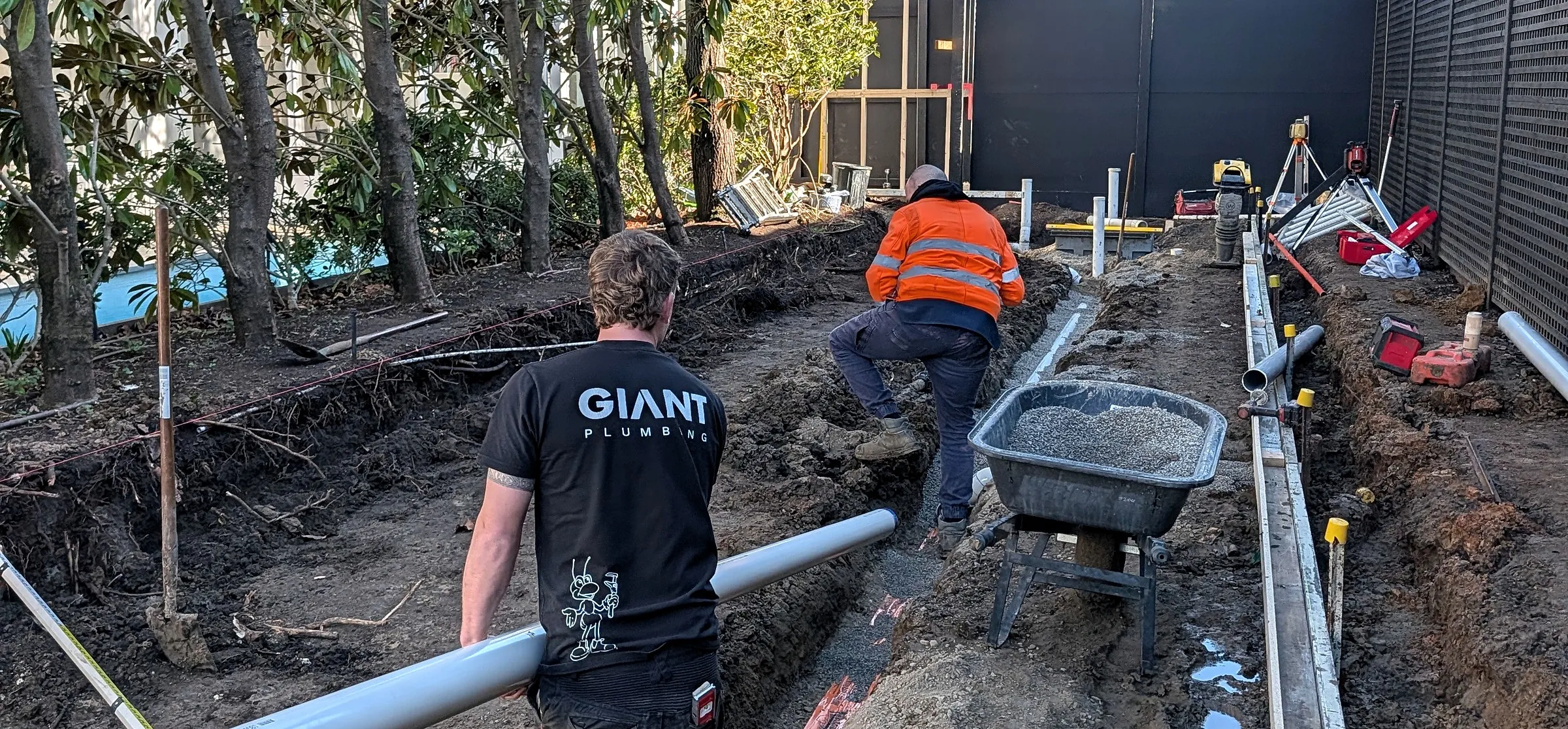 Two Giant Plumbing team members lay new drainage in a long trench beside a house