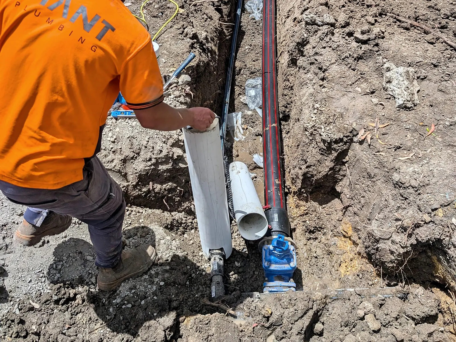 A Giant Plumbing team member lowers new pipe beside a blue valve on a water main in an open trench