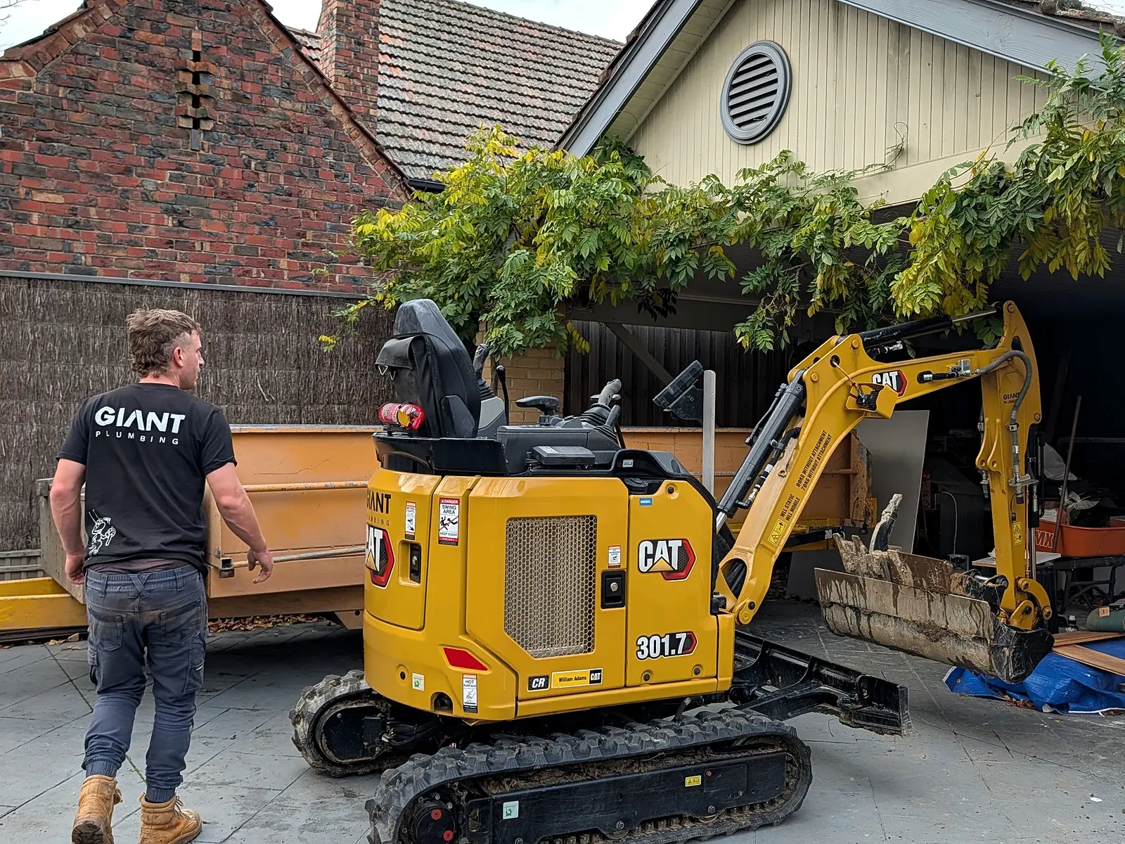 Giant Plumbing's smallest excavator at work in a paved courtyard beside a tipping trailer, a team member walking past