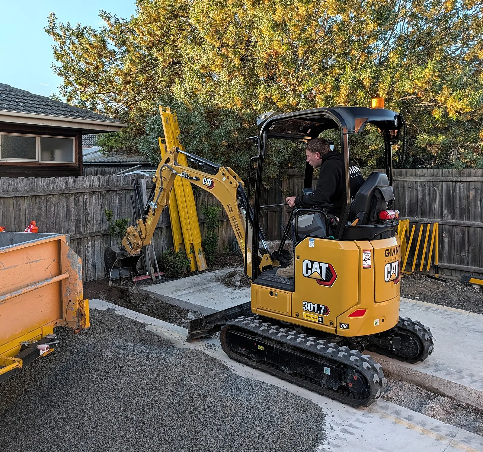 A Giant Plumbing operator in the cab of the company's mini excavator in a backyard, the tipping trailer beside it