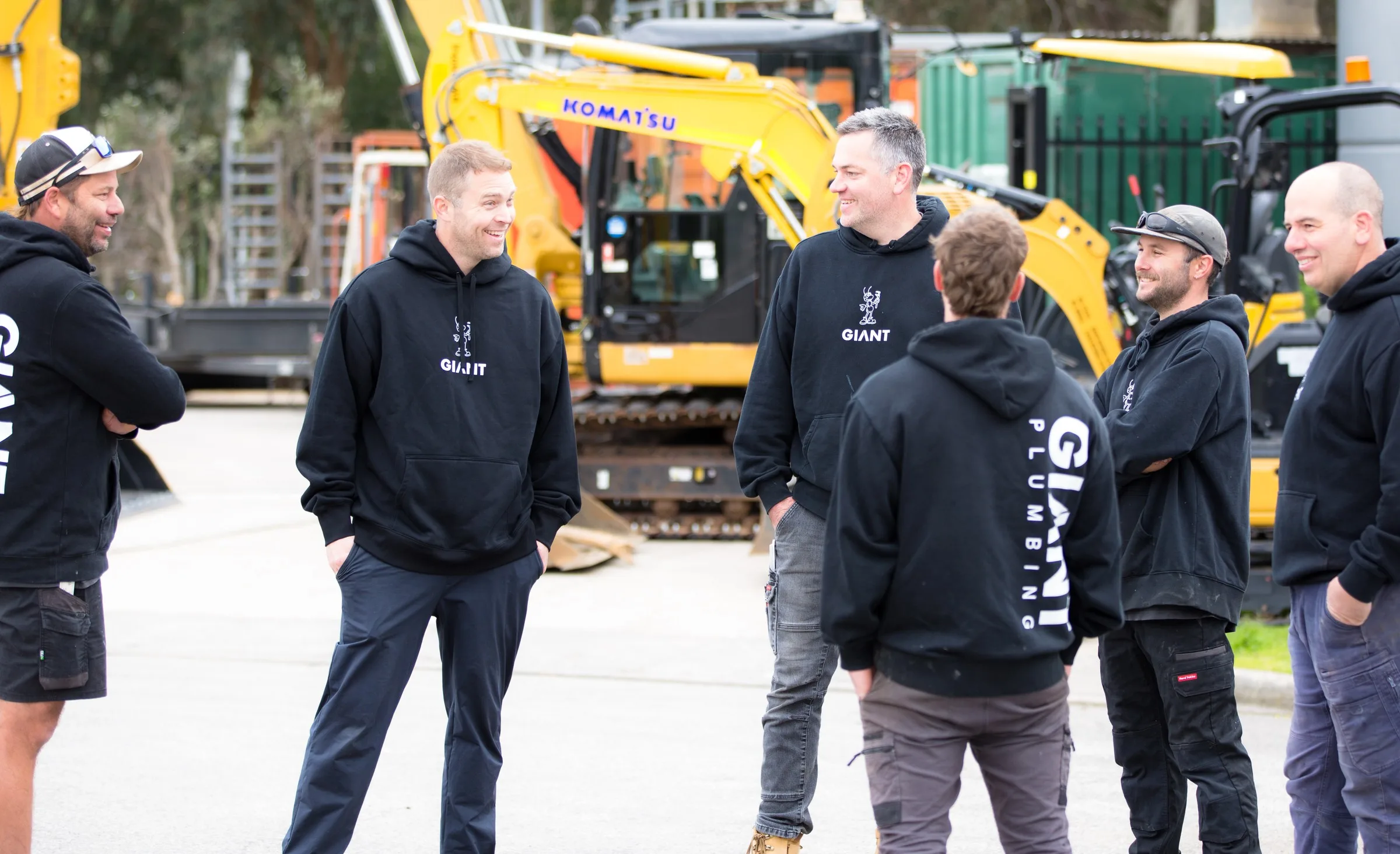 Six of the Giant Plumbing team in GIANT hoodies talking at the Coburg North yard, their excavators behind them