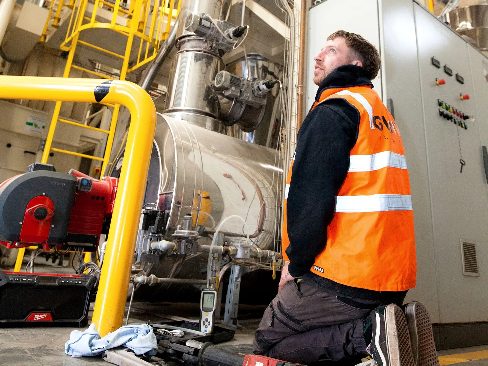A Giant Plumbing technician in hi-vis kneels beside an industrial burner and checks the plant above