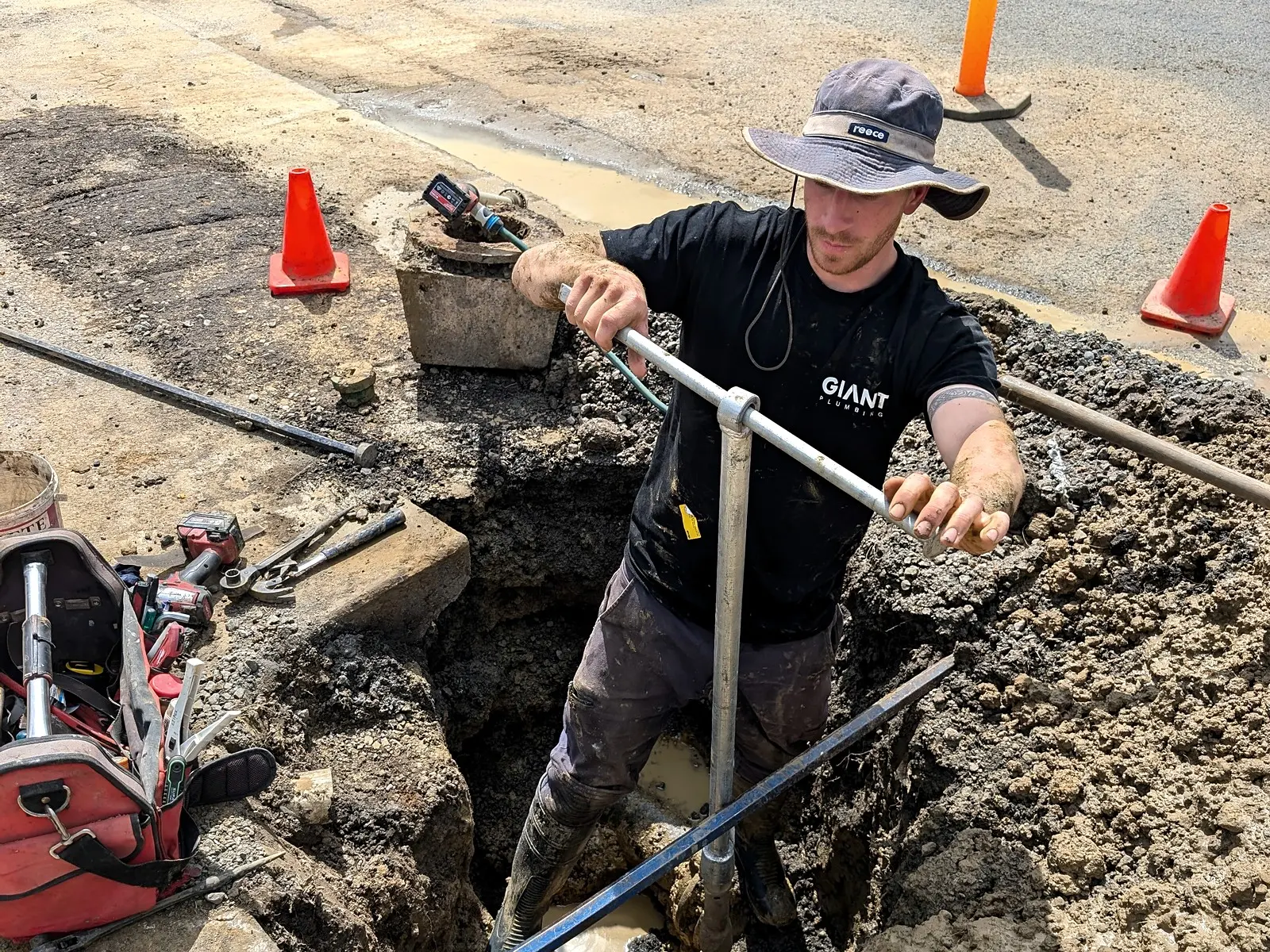 A Giant Plumbing team member repairs a burst water service in a road excavation, traffic cones around him