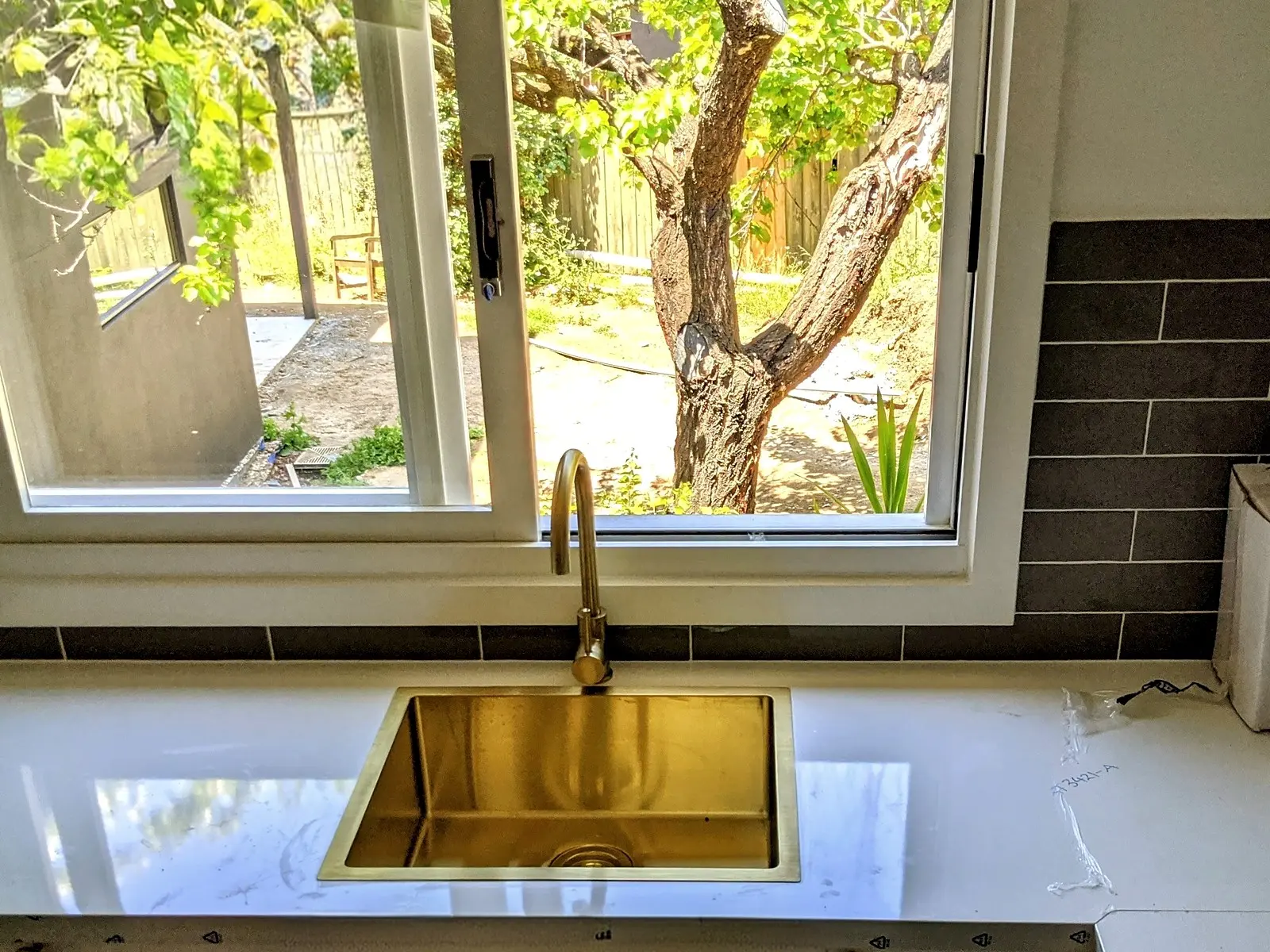 A brass mixer tap and sink set into a stone benchtop under a kitchen window