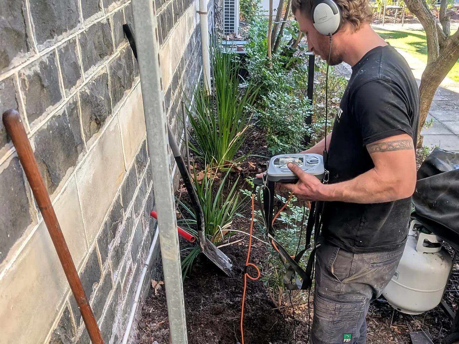 A Giant Plumbing team member listens for a leak with a ground microphone and headphones beside a bluestone wall