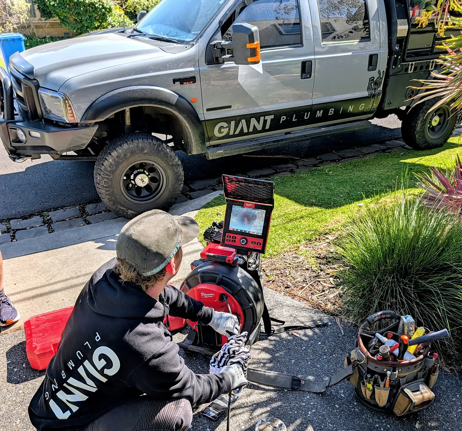 A Giant Plumbing team member runs the drain camera into a driveway drain, the monitor on the reel and the GIANT PLUMBING truck behind him