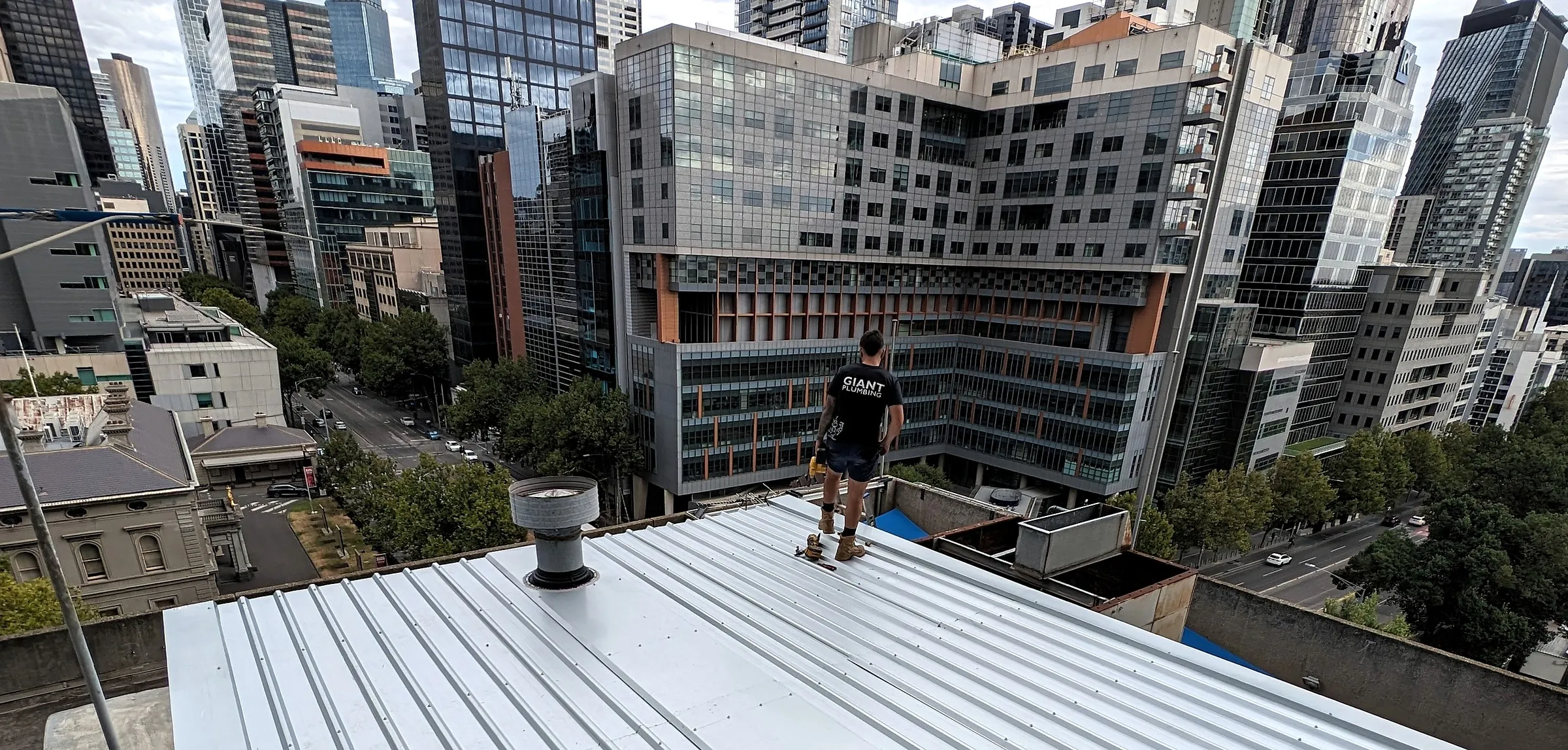 A Giant Plumbing team member stands on a new metal roof on a city rooftop, the Melbourne skyline behind