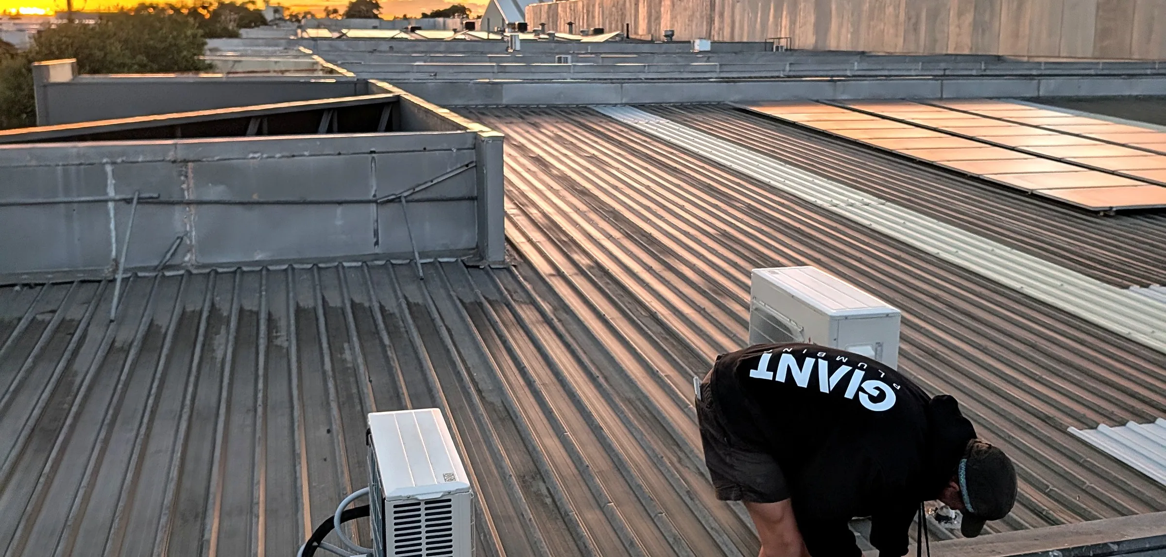 A Giant Plumbing team member works at the box gutter edge of a commercial roof at sunrise, air conditioning units and solar panels beside him