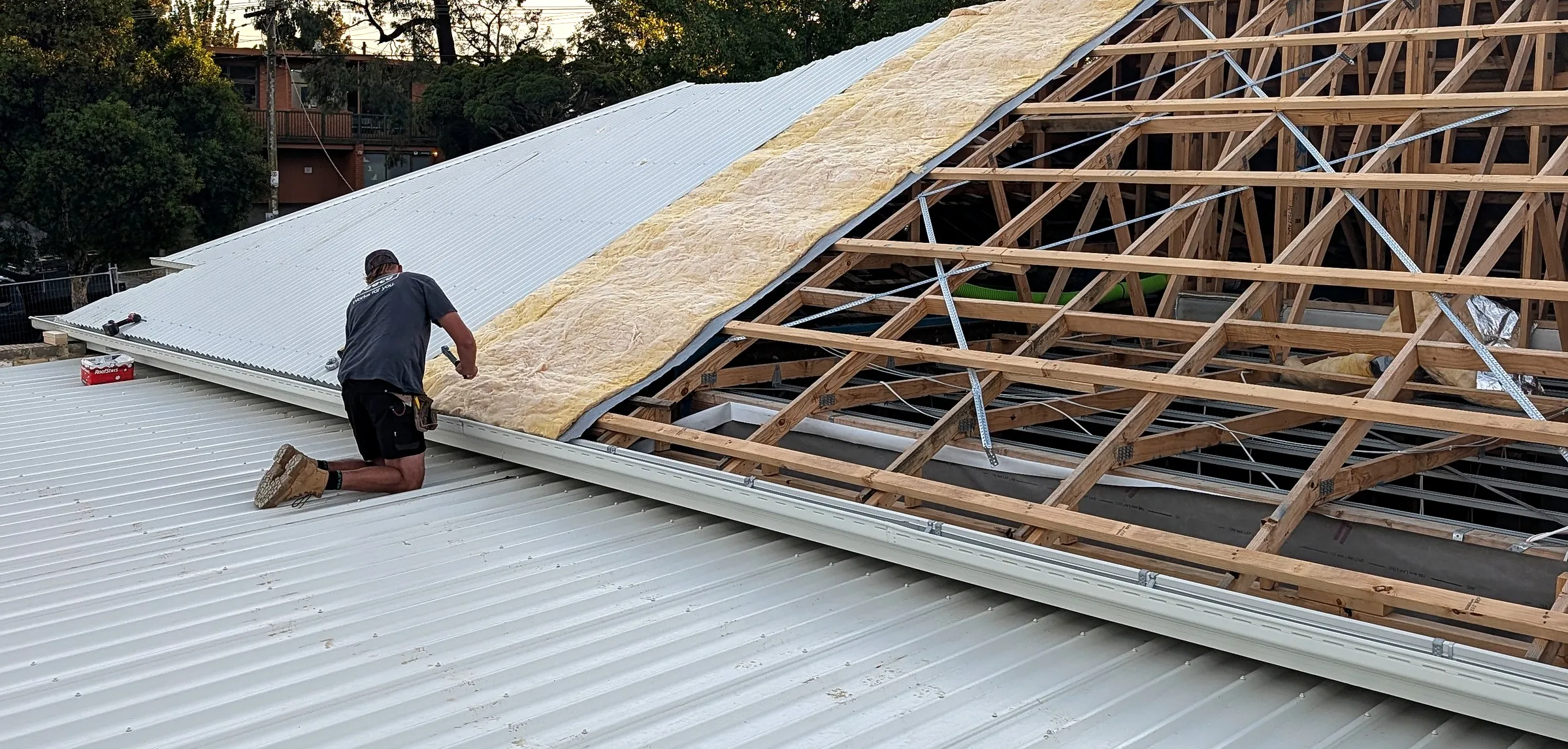 New metal roof sheets going on over the stripped trusses and insulation of a house at dusk, two roofers at work