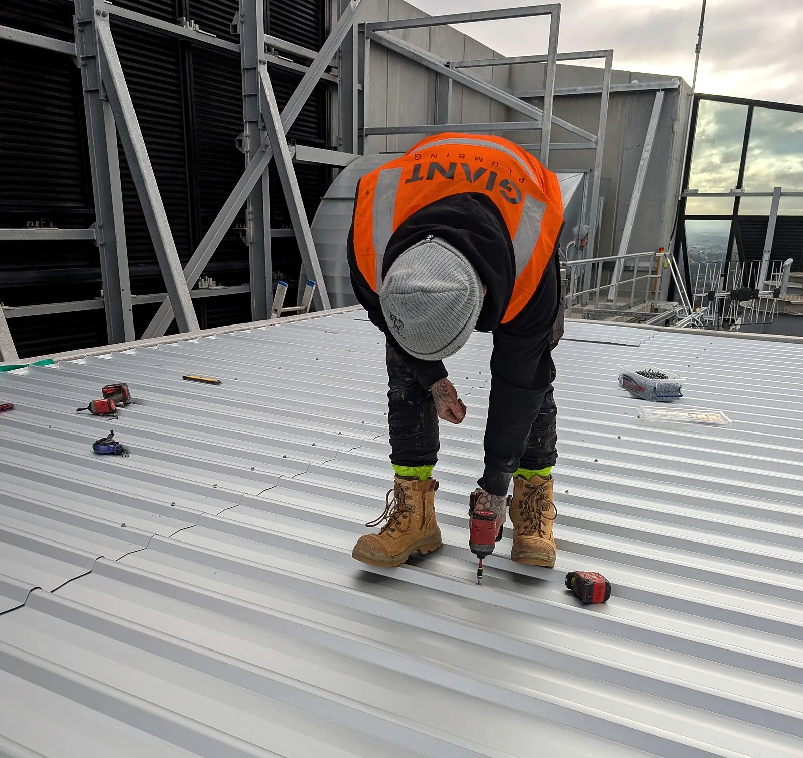 A Giant Plumbing roofer in a branded hi-vis vest screws down new roof sheets on top of a high-rise building